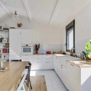 Aménagement d'une cuisine américaine contemporaine en L avec un évier de ferme, un placard à porte plane, des portes de placard blanches, un plan de travail en bois, une crédence blanche, une crédence en carreau de verre, un électroménager blanc, un sol blanc et un plan de travail beige.