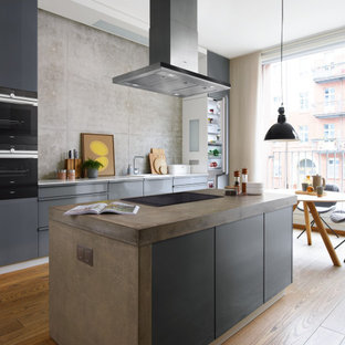 Cette image montre une cuisine ouverte parallèle design avec des portes de placard grises, un plan de travail en béton, une crédence grise, un électroménager encastrable, un sol en bois brun, un îlot central, un sol marron et un plan de travail gris.