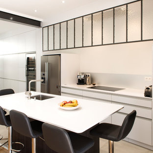 Cette image montre une cuisine design avec un évier 1 bac, des portes de placard blanches, un plan de travail en surface solide, une crédence blanche, un électroménager en acier inoxydable, un îlot central, un placard à porte plane et un sol beige.