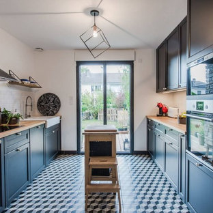 Cette image montre une cuisine parallèle traditionnelle de taille moyenne avec un évier de ferme, des portes de placard noires, un plan de travail en bois, une crédence en carrelage métro, un sol en carreaux de ciment, un îlot central, un placard avec porte à panneau encastré, une crédence blanche, un électroménager en acier inoxydable, un sol multicolore et un plan de travail marron.
