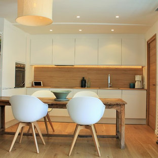 Cette image montre une cuisine américaine design en L avec un placard à porte plane, des portes de placard blanches, une crédence marron, un sol en bois brun et un sol marron.