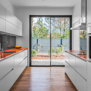 Cette image montre une cuisine parallèle design avec un évier encastré, un placard à porte plane, des portes de placard blanches, un plan de travail en verre, une crédence grise, une crédence en feuille de verre, un électroménager en acier inoxydable, un sol en bois brun, aucun îlot et un plan de travail orange.