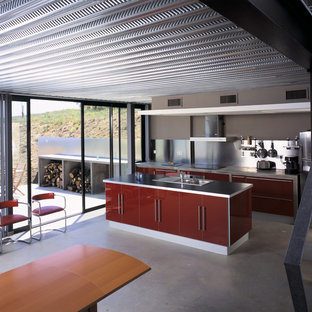 Idées déco pour une cuisine américaine parallèle industrielle de taille moyenne avec un placard à porte plane, des portes de placard rouges, un plan de travail en béton, une crédence métallisée, un électroménager en acier inoxydable, béton au sol et un îlot central.