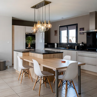 Cette image montre une cuisine américaine design en L avec un placard à porte plane, des portes de placard blanches, une crédence noire, un électroménager encastrable, un îlot central, un sol beige et un plan de travail noir.