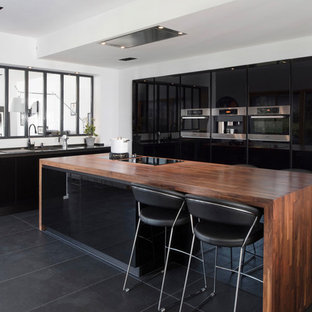 Cette photo montre une cuisine tendance avec un évier encastré, un placard à porte plane, des portes de placard noires, un plan de travail en bois, une crédence en fenêtre, un électroménager en acier inoxydable, un îlot central et un sol noir.