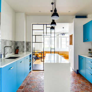 Inspiration pour une cuisine parallèle design fermée avec un évier encastré, un placard à porte plane, des portes de placard bleues, une crédence verte, un sol en brique, un îlot central, un sol rouge et un plan de travail blanc.