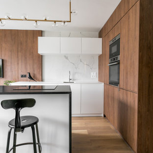Exemple d'une grand cuisine américaine tendance en L avec un évier encastré, un placard à porte plane, des portes de placard blanches, un plan de travail en granite, une crédence blanche, une crédence en marbre, un électroménager noir, un sol en bois clair, un îlot central, un sol marron et un plan de travail noir.
