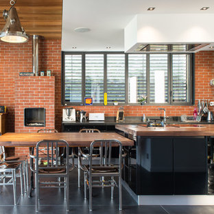 Cette photo montre une grand cuisine américaine parallèle industrielle avec un placard à porte plane, des portes de placard en bois sombre, un plan de travail en bois, une crédence rouge, un électroménager noir, béton au sol et un îlot central.