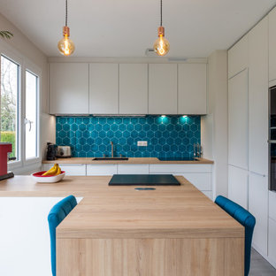 Cette photo montre une cuisine tendance en U de taille moyenne avec un évier encastré, un placard à porte plane, des portes de placard blanches, un plan de travail en bois, une crédence bleue, une crédence en carreau de porcelaine, un électroménager encastrable, un sol en carrelage de porcelaine, une péninsule, un sol gris et un plan de travail beige.