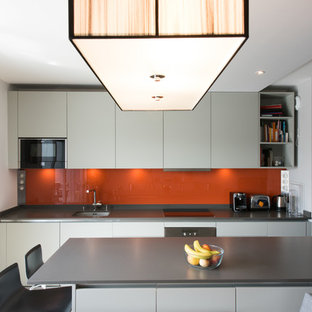 Cette photo montre une cuisine américaine parallèle tendance de taille moyenne avec une crédence orange, une crédence en feuille de verre, un îlot central, un évier encastré, un placard à porte plane, des portes de placard blanches et un électroménager encastrable.