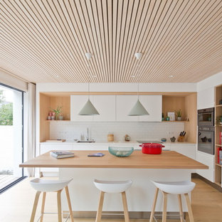 Idées déco pour une cuisine américaine bord de mer en L de taille moyenne avec un évier encastré, des portes de placard blanches, un plan de travail en quartz modifié, une crédence blanche, un électroménager encastrable, un sol en bois clair, un îlot central, un plan de travail blanc, un placard à porte plane, une crédence en carrelage métro et un sol marron.