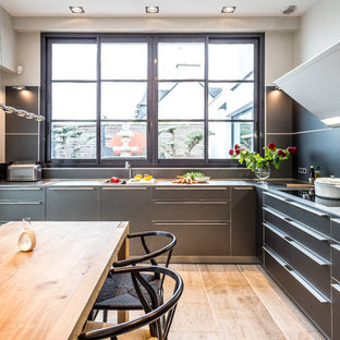 Cette image montre une cuisine américaine design en L de taille moyenne avec un placard à porte plane, des portes de placard grises, aucun îlot, un évier 1 bac, un plan de travail en inox, une crédence noire, un électroménager encastrable, un sol en bois brun, un sol marron et un plan de travail gris.