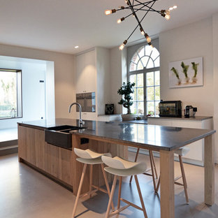 Réalisation d'une grand cuisine américaine parallèle design avec un évier de ferme, un îlot central, un électroménager encastrable, un placard à porte affleurante, des portes de placard en bois clair, un plan de travail en granite, une crédence noire, une crédence en carreau de terre cuite et béton au sol.