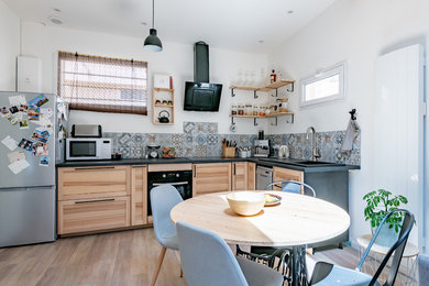 This is an example of a medium sized contemporary l-shaped open plan kitchen in Bordeaux with a single-bowl sink, flat-panel cabinets, light wood cabinets, granite worktops, blue splashback, ceramic splashback, black appliances, light hardwood flooring, no island, brown floors and black worktops.