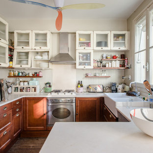 Exemple d'une cuisine éclectique en U avec une péninsule, un évier de ferme, un placard à porte vitrée, un électroménager en acier inoxydable, un sol en bois foncé, un sol marron, des portes de placard beiges, une crédence beige et un plan de travail beige.