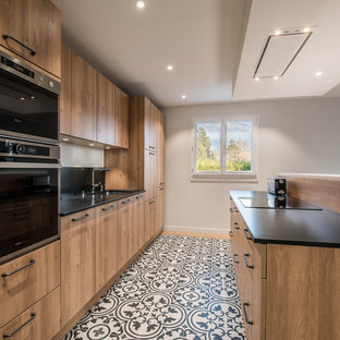 Cette photo montre une cuisine ouverte parallèle tendance de taille moyenne avec un évier encastré, un placard à porte plane, des portes de placard en bois brun, un électroménager noir, un sol en carreaux de ciment, un îlot central et une crédence noire.