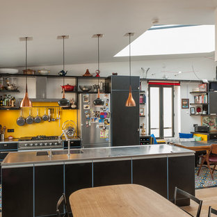 Exemple d'une grand cuisine ouverte parallèle éclectique avec des portes de placard noires, un îlot central, un évier intégré, une crédence jaune, un électroménager noir et un plan de travail gris.