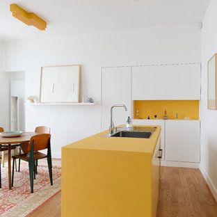 Inspiration pour une petit cuisine américaine linéaire design avec un évier 1 bac, un placard à porte plane, des portes de placard blanches, une crédence jaune, un sol en bois clair, un îlot central et un plan de travail jaune.