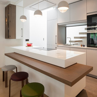 Réalisation d'une cuisine design avec un évier encastré, un placard à porte plane, des portes de placard blanches, une crédence en fenêtre, un électroménager noir, une péninsule, un sol marron et un plan de travail blanc.