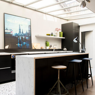 Aménagement d'une cuisine parallèle contemporaine avec un placard à porte plane, des portes de placard noires, un sol en bois clair, un îlot central, un sol beige et un plan de travail blanc.