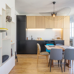 Cette photo montre une petit cuisine américaine linéaire rétro avec un évier encastré, une crédence blanche, un électroménager encastrable, un placard à porte plane, des portes de placard noires, un sol en bois brun, aucun îlot et un sol marron.