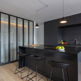Cette photo montre une cuisine ouverte parallèle tendance de taille moyenne avec un évier intégré, un placard à porte plane, des portes de placard noires, un plan de travail en granite, une crédence noire, un électroménager en acier inoxydable, un sol en bois clair et un îlot central.