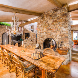 Idee per un'ampia sala da pranzo aperta verso la cucina mediterranea con pareti marroni, pavimento in cemento, camino bifacciale e cornice del camino in pietra