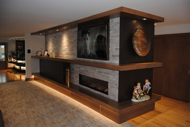 Imagen de sala de estar con barra de bar abierta contemporánea de tamaño medio con suelo de madera clara, chimenea lineal, marco de chimenea de piedra, televisor colgado en la pared, paredes grises y suelo marrón