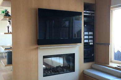Modelo de sala de estar cerrada contemporánea de tamaño medio con paredes beige, suelo de madera clara, chimenea de doble cara, marco de chimenea de yeso, televisor colgado en la pared y suelo marrón