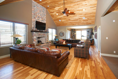 Modelo de sala de estar abierta de estilo americano con paredes beige, suelo de madera en tonos medios, todas las chimeneas, marco de chimenea de piedra y televisor colgado en la pared