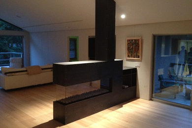 Imagen de sala de estar con barra de bar cerrada contemporánea de tamaño medio con suelo de madera clara, chimenea de doble cara, marco de chimenea de metal y pared multimedia