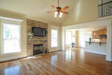 Imagen de sala de estar abierta grande con paredes beige, suelo de madera clara, todas las chimeneas, marco de chimenea de piedra y televisor colgado en la pared