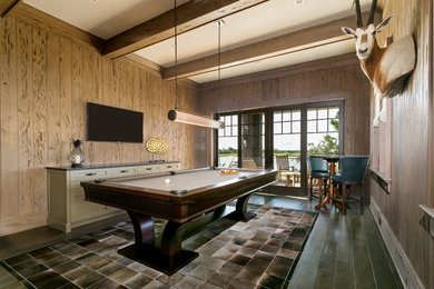 Modelo de sala de juegos en casa costera grande con paredes beige, suelo de madera oscura, televisor colgado en la pared y suelo marrón