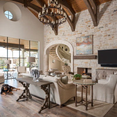 Huge tuscan open concept brown floor and medium tone wood floor family room photo in Dallas with a standard fireplace, a stone fireplace, a wall-mounted tv and beige walls
