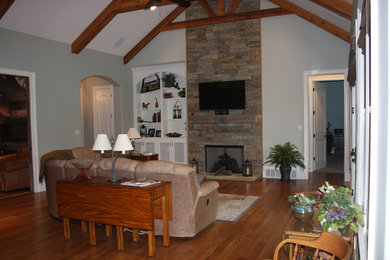Modelo de sala de estar abierta grande con paredes grises, suelo de madera en tonos medios, todas las chimeneas, marco de chimenea de piedra y televisor colgado en la pared