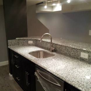 Exemple d'un très grand bar de salon avec évier linéaire tendance avec un placard à porte shaker, des portes de placard en bois sombre, un plan de travail en granite et un sol en bois clair.
