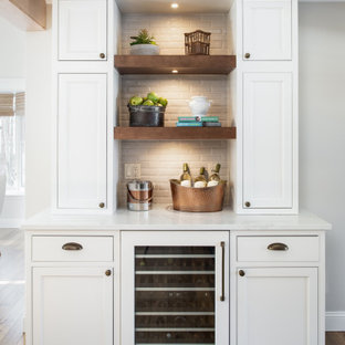 Exemple d'un bar de salon chic de taille moyenne avec un évier encastré, un placard à porte affleurante, des portes de placard blanches, un plan de travail en quartz modifié, une crédence beige, une crédence en carreau de porcelaine, un sol en bois clair et un plan de travail blanc.