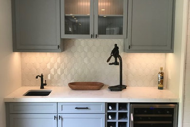 Example of a mid-sized minimalist single-wall medium tone wood floor and brown floor wet bar design in Other with an undermount sink, recessed-panel cabinets, gray cabinets, quartzite countertops, gray backsplash, ceramic backsplash and white countertops