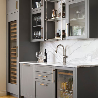 Imagen de bar en casa con fregadero lineal, tradicional renovado, con fregadero bajoencimera, armarios tipo vitrina, puertas de armario grises, salpicadero blanco, suelo de madera clara, suelo beige, encimera de mármol, salpicadero de mármol y encimeras blancas