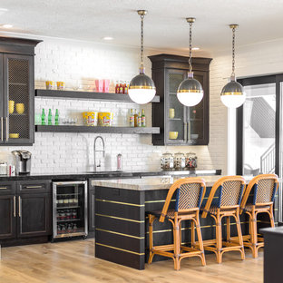 Foto de bar en casa con barra de bar tradicional renovado, grande, con armarios estilo shaker, salpicadero blanco, suelo de madera clara, fregadero bajoencimera, puertas de armario negras, encimera de mármol, salpicadero de azulejos tipo metro, suelo marrón y encimeras blancas