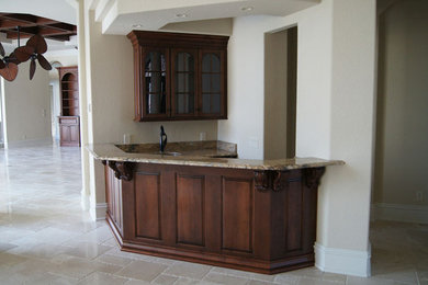 Example of a mid-sized classic single-wall travertine floor and beige floor wet bar design in Charlotte with an undermount sink, raised-panel cabinets, dark wood cabinets, granite countertops, beige backsplash and stone slab backsplash