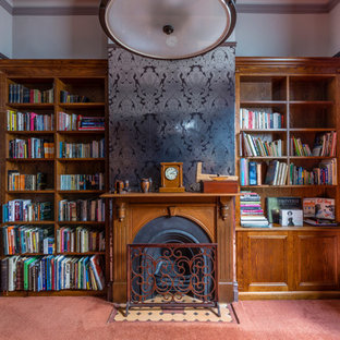 Bild på ett mellanstort vintage hemmabibliotek, med grå väggar, heltäckningsmatta, en standard öppen spis, en spiselkrans i trä, ett fristående skrivbord och orange golv