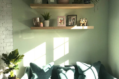 Modelo de despacho escandinavo pequeño con paredes verdes, suelo de madera en tonos medios, chimenea de esquina, marco de chimenea de ladrillo y escritorio empotrado