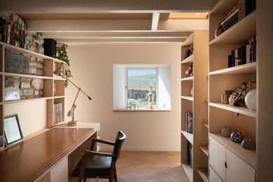 Modelo de despacho escandinavo pequeño sin chimenea con paredes blancas, suelo de madera clara, escritorio empotrado, suelo marrón y vigas vistas