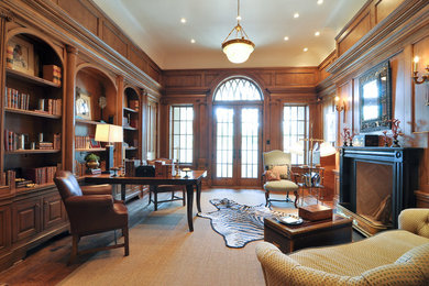 Klassisk inredning av ett stort hemmabibliotek, med bruna väggar, mellanmörkt trägolv, en standard öppen spis, ett fristående skrivbord och en spiselkrans i trä