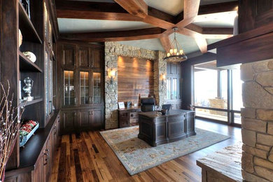 This is an example of a rustic home office in Other with a two-sided fireplace, a stone fireplace surround, a freestanding desk and brown floors.