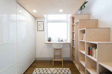 Diseño de despacho escandinavo sin chimenea con biblioteca, paredes blancas, suelo de madera oscura, escritorio empotrado y suelo marrón