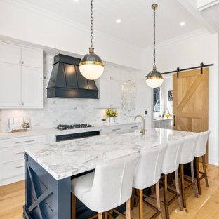 This is an example of a country galley kitchen in Brisbane with a farmhouse sink, shaker cabinets, white cabinets, white splashback, stainless steel appliances, medium hardwood floors, with island, brown floor and white benchtop.