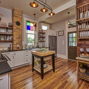 Kitchen - mid-sized eclectic l-shaped medium tone wood floor and brown floor kitchen idea in Other with a farmhouse sink, shaker cabinets, white cabinets, granite countertops, white backsplash, stainless steel appliances, an island, black countertops and subway tile backsplash