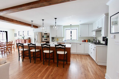 Design ideas for an expansive farmhouse l-shaped open plan kitchen in New York with a submerged sink, shaker cabinets, white cabinets, engineered stone countertops, beige splashback, ceramic splashback, stainless steel appliances, medium hardwood flooring, an island, brown floors and white worktops.